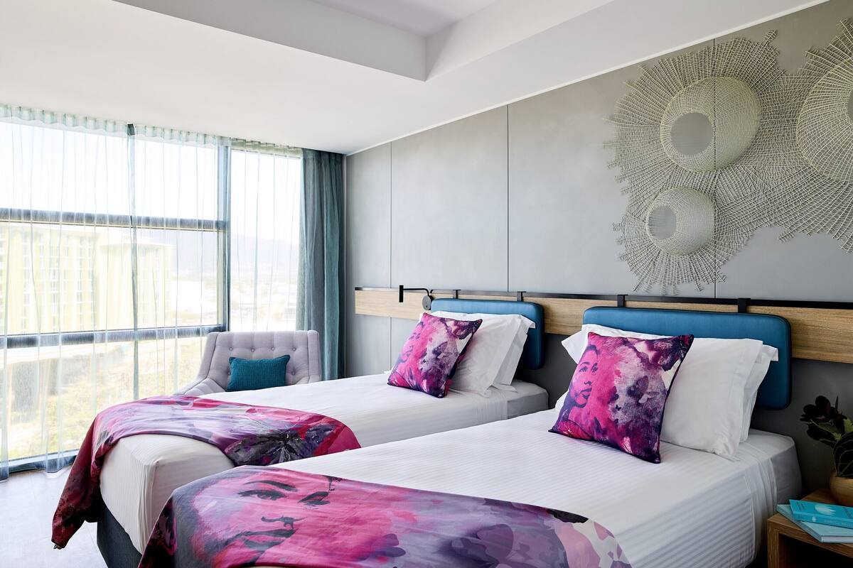 A modern hotel room with two beds, a large window, and a decorative wall piece.