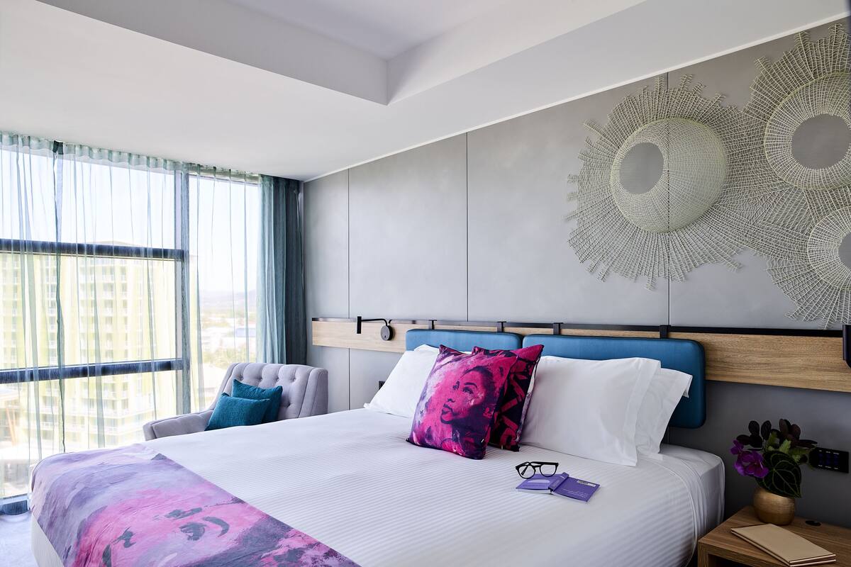 A modern hotel room with a large bed, a chair, a bedside table with a plant, and a view of the city through large windows.