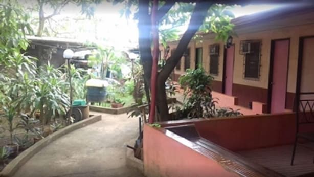 A covered outdoor area with a concrete path leading to a building with pink walls and windows. There are potted plants and a small table with a chair. - EG Rusiana Boarding House (Claver)