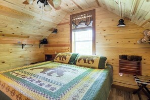 Una habitación acogedora con techo de madera, una cama con colcha, una ventana con cortinas, un estante con un osito de peluche y una mesita.