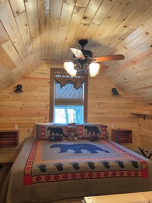 A wooden cabin bedroom with a bed, a ceiling fan, a window, and a bear-themed bedspread.