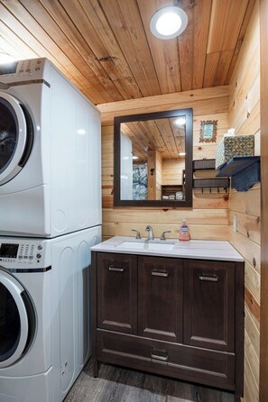 A compact bathroom with a washing machine, a sink with two faucets, and a mirror above the vanity. - Sleepin Bear Treehouse - Private Hot Tub (Guild)