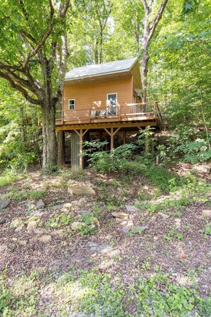 A wooden cabin with a deck, surrounded by trees and natural ground cover. - Sleepin Bear Treehouse - Private Hot Tub (Guild)