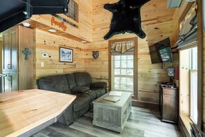 A cabin interior with a wooden interior, a sofa, a small table, a mounted television, and a mounted animal hide. - Sleepin Bear Treehouse - Private Hot Tub (Guild)