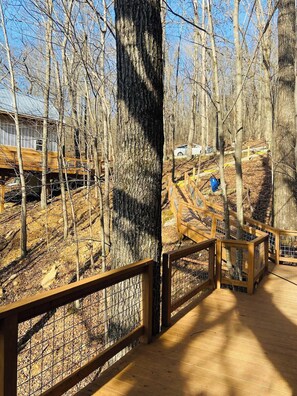 A wooden deck with a metal railing, surrounded by trees and a building in the background. - Sleepin Bear Treehouse - Private Hot Tub (Guild)