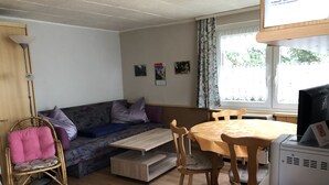 Ein gemütliches Wohnzimmer mit einer Couch, einem Esstisch und Stühlen. Es gibt eine Lampe, einen Drucker und ein Fenster mit Vorhängen.