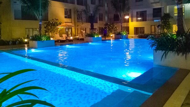Une piscine nocturne, dotée de chaises longues et bordée de palmiers, entourée d’un immeuble à plusieurs étages.