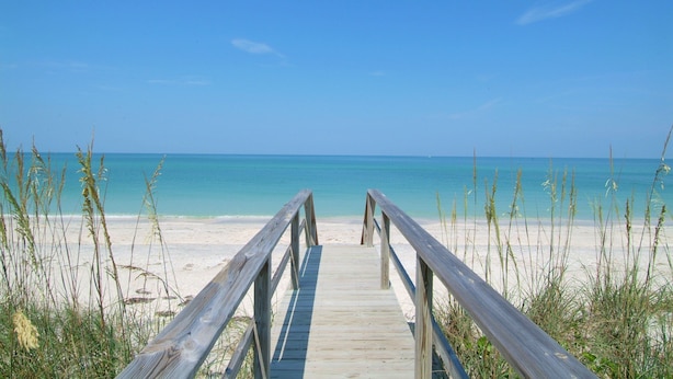Un paseo marítimo de madera que conduce a una playa con aguas azul cristalino y orilla arenosa.