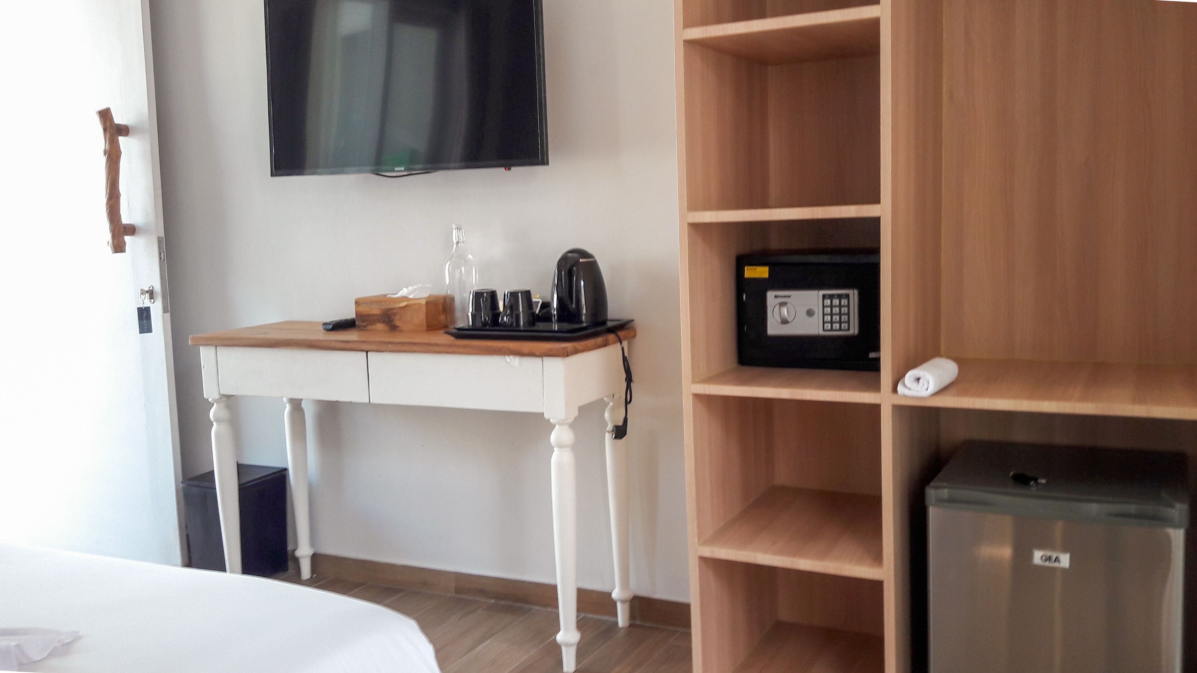 A hotel room with a wooden desk, a television mounted on the wall, a minibar, and a bed.