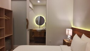 A hotel room with a bed, a bedside table with a lamp, a wooden wardrobe, a round mirror with a light, and a sink. - Mantra Gili (Gili Trawangan)