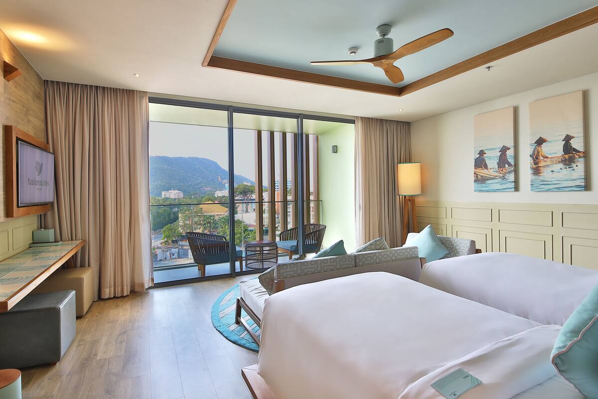 Habitación de hotel con una cama grande, un escritorio, una silla y un balcón con vistas a la ciudad y las montañas.