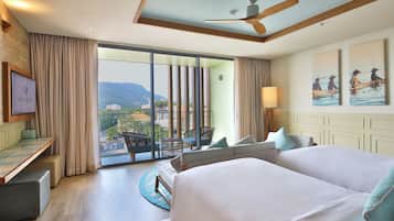 A hotel room with a large bed, a desk, a chair, and a balcony with a view of the city and mountains.