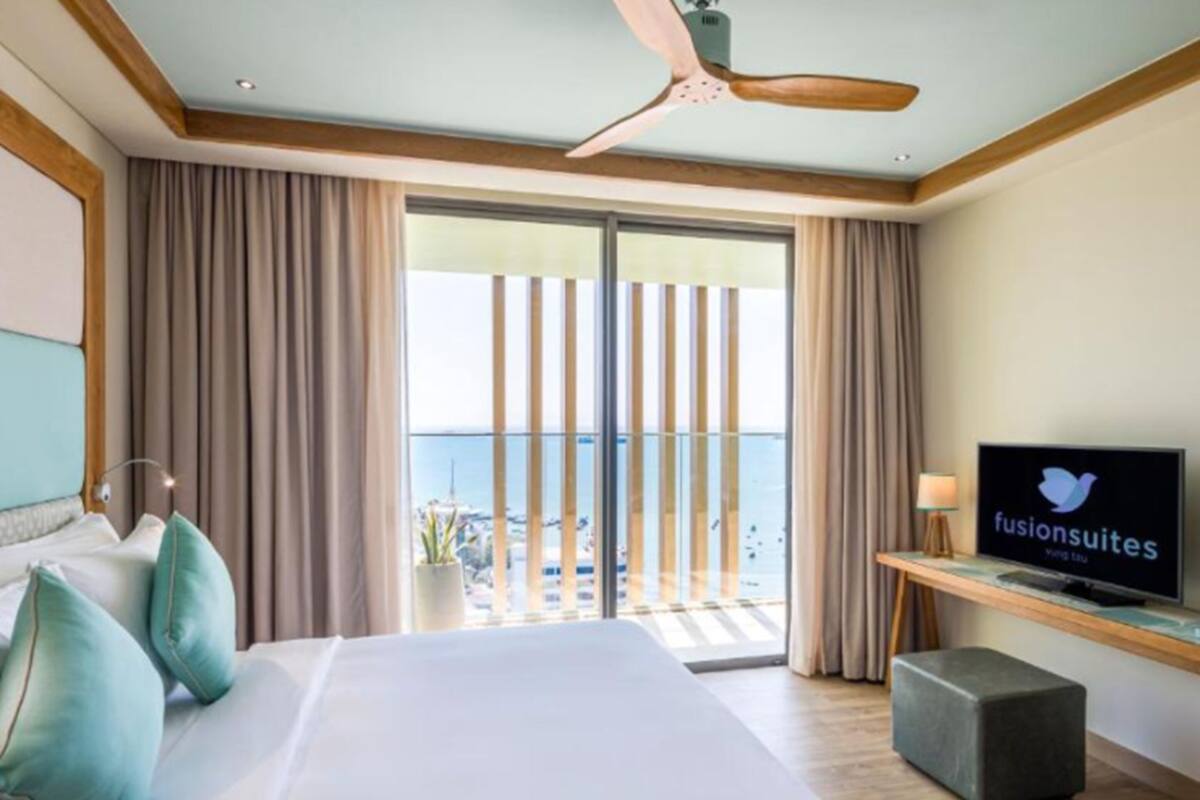 Habitación de hotel con una cama grande, televisor, balcón con vista al mar y ventilador de techo.