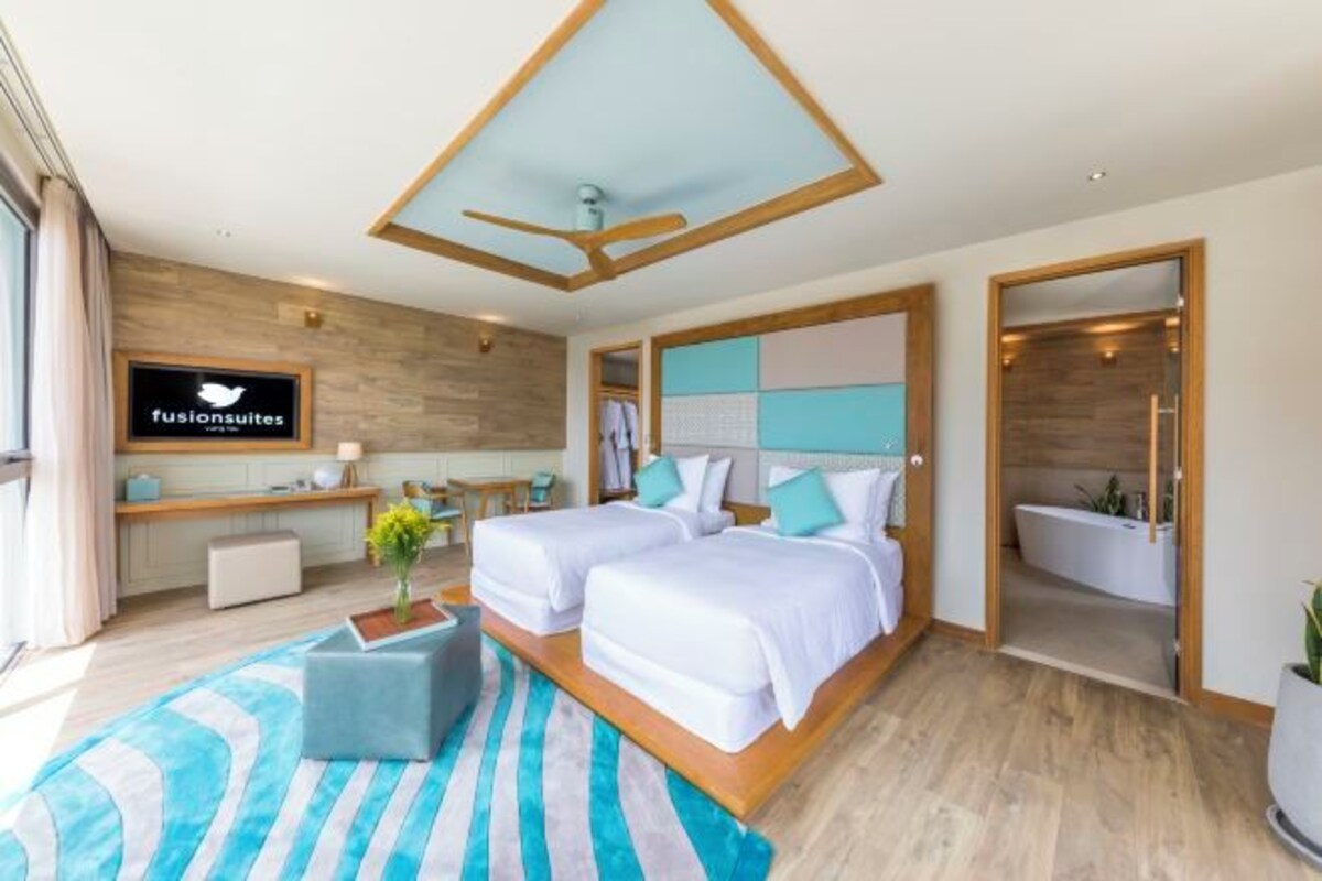 A modern hotel room with two beds, a ceiling fan, a TV, a bathroom, and a wooden floor.