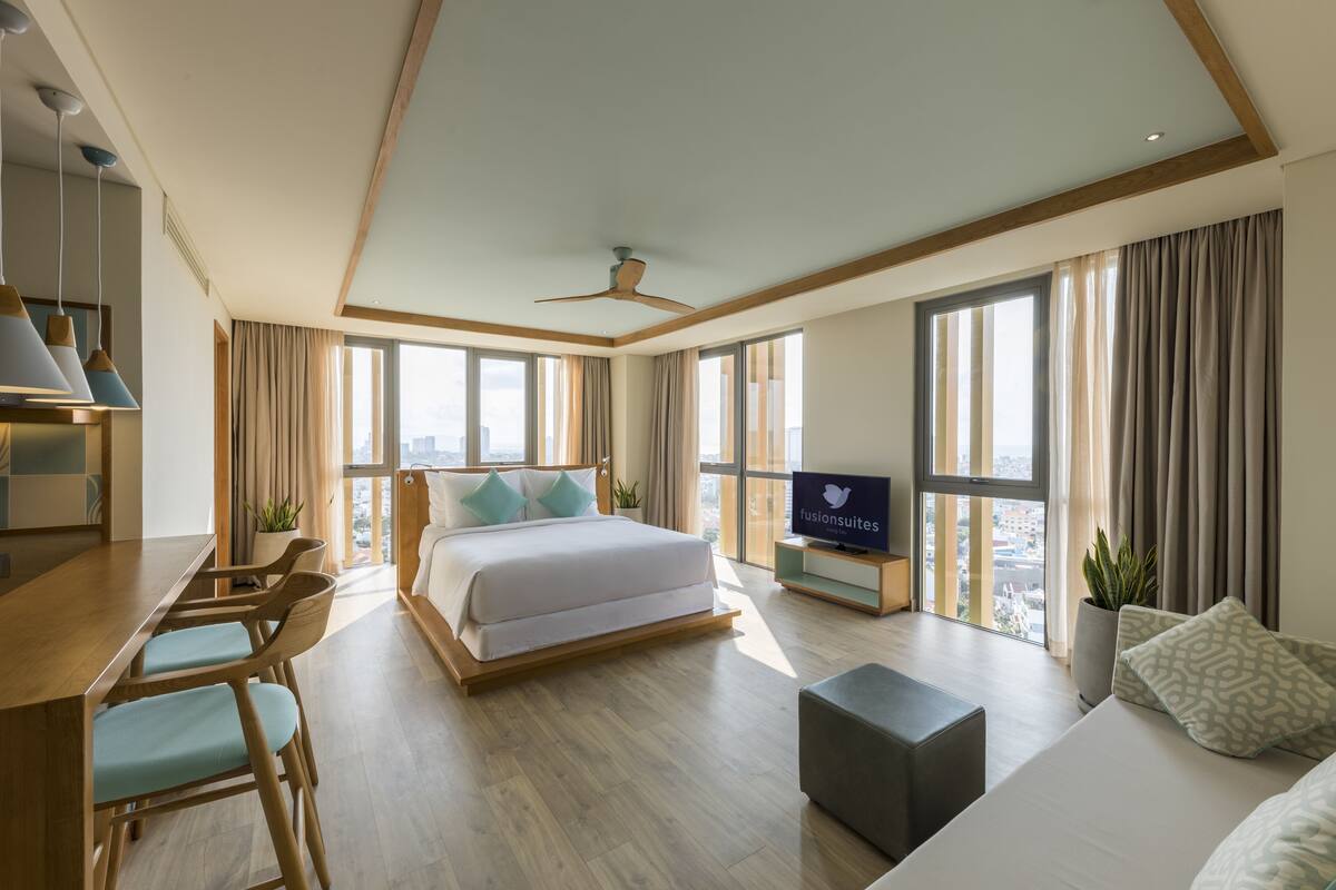 Una habitación de hotel moderna con una cama grande, un escritorio con sillas, un televisor y ventanas amplias con vistas a la ciudad.