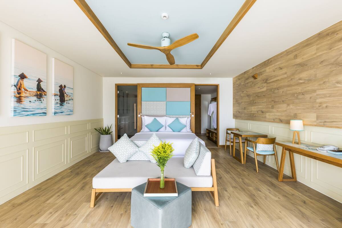 Una habitación de hotel moderna con ventilador de techo de madera, una cama grande con almohadas estampadas, un área de descanso con una mesita auxiliar y una pared de madera.