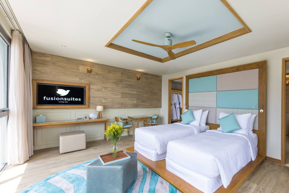 Habitación de hotel moderna con techo de madera, dos camas, un escritorio y un televisor mostrando 'fusionsuites'.