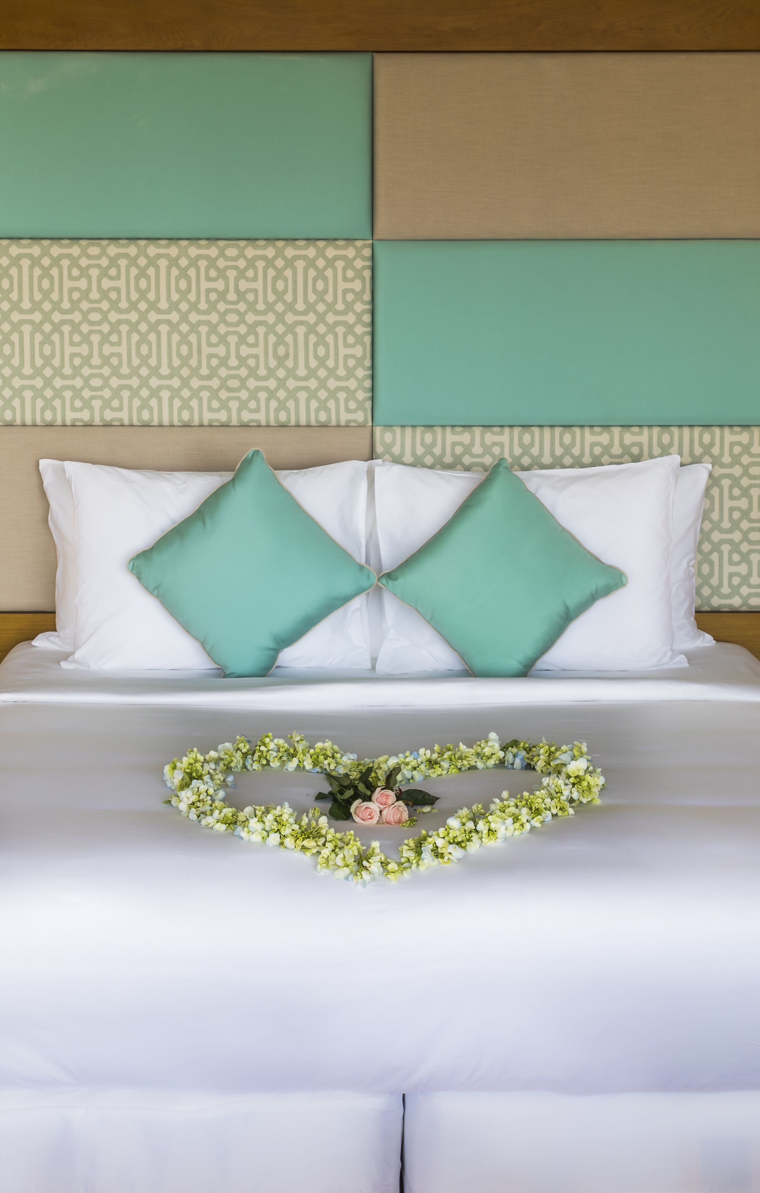 A neatly made bed with white bedding and two teal pillows, a floral garland on the bed, and a patterned headboard.