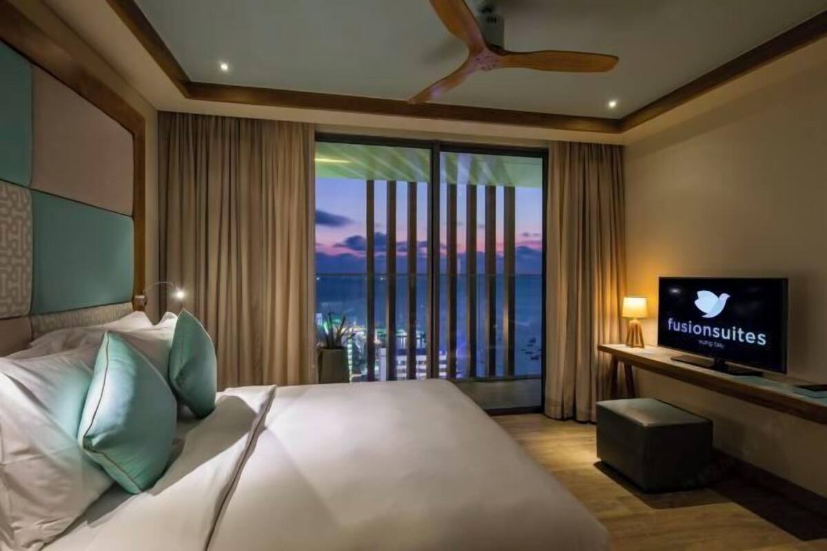 Habitación de hotel con cama, escritorio con televisor, ventilador de techo y vista a la ciudad al anochecer.
