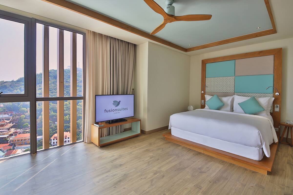 Una habitación de hotel con un ventanal grande, una cama, un televisor sobre un soporte de madera y un ventilador de techo.