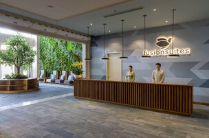 A reception area with a wooden reception desk, two receptionists, a large potted plant, and a wall with a geometric pattern and a logo.