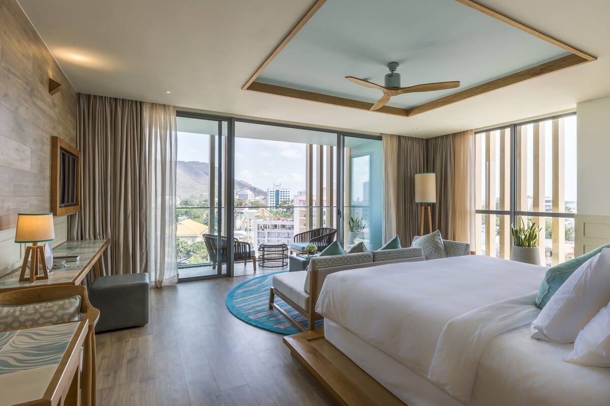 Habitación de hotel con una cama grande, un escritorio, una silla y vistas a la ciudad a través de amplias ventanas.