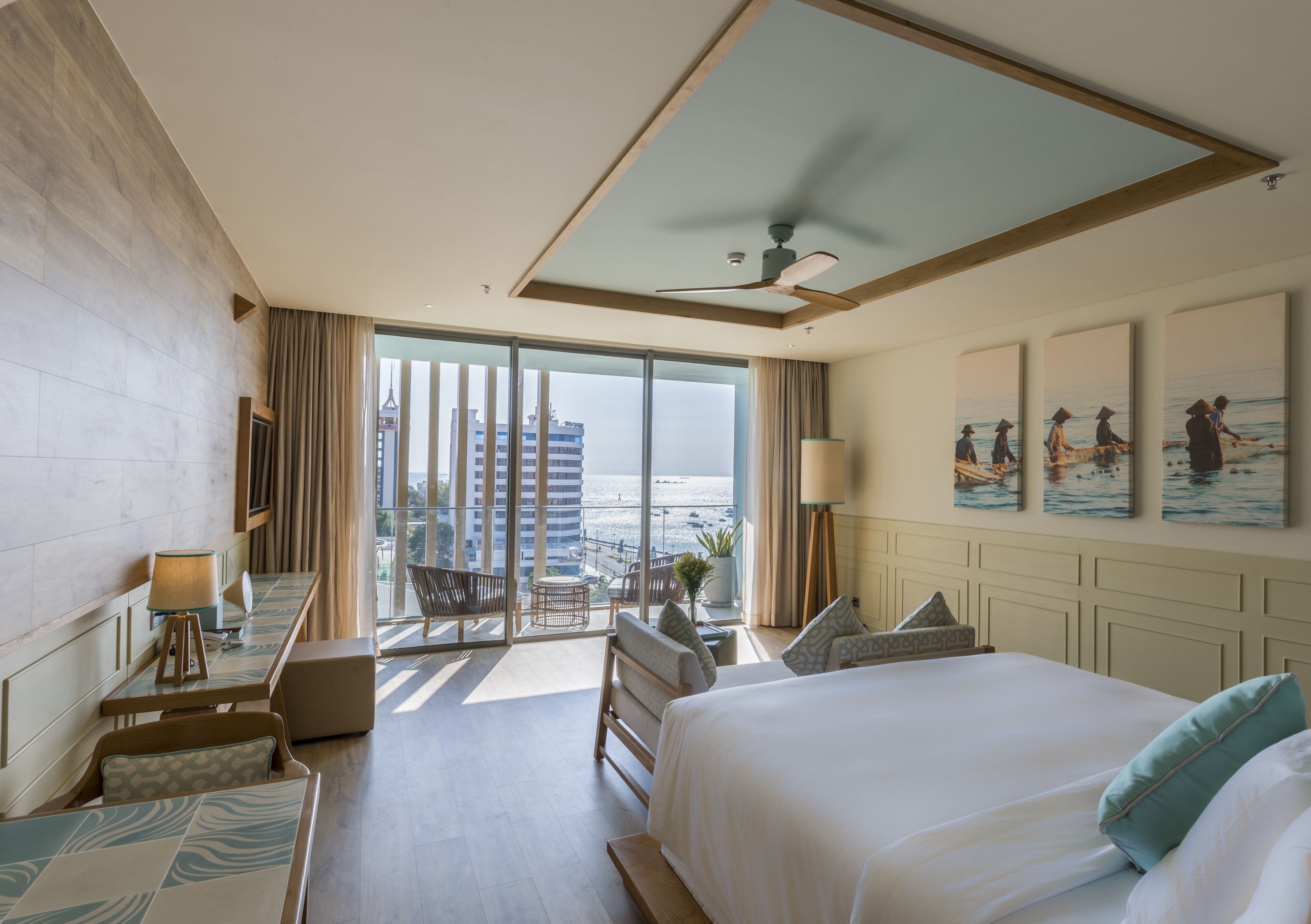 A hotel room with a large bed, a desk, a chair, a balcony with a view of the city, and artwork on the walls.