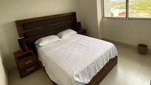 Ein Schlafzimmer mit einem hölzernen Bett, weißer Bettwäsche, einem Nachttisch, einer Lampe und einem Fenster mit Blick auf eine Stadtlandschaft.