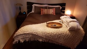 A neatly made bed with a fluffy white throw, a wooden tray with a book and a remote, and a white towel on the side.