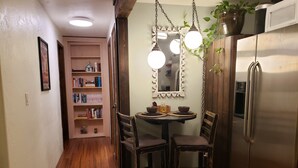 A kitchen with wooden flooring, a refrigerator, a dining table with chairs, and a bookshelf.