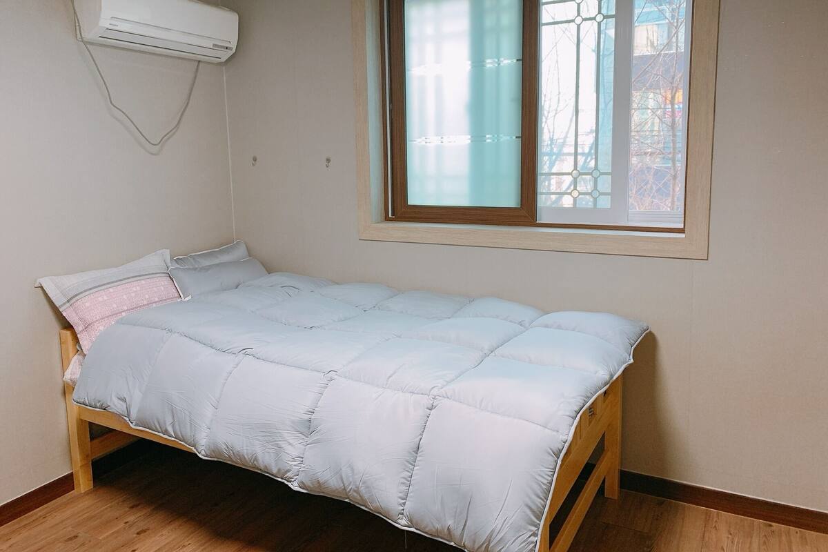 Un lit simple avec une couette blanche, dans une pièce dotée d’une fenêtre et d’un climatiseur.