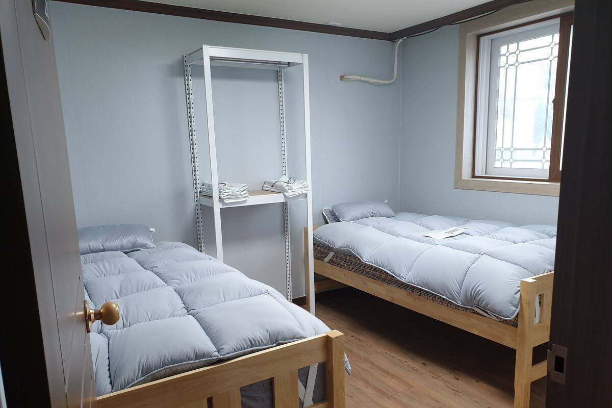 Une pièce avec deux lits en bois, une étagère en métal blanche et une fenêtre avec des stores.