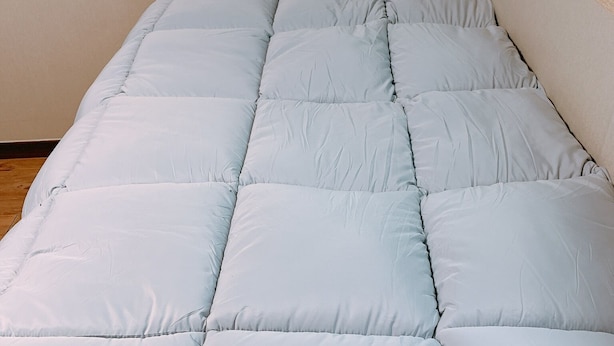 Een bed met een lichtblauwe quilt en kussens, een houten vloer en een raam met houten kader.