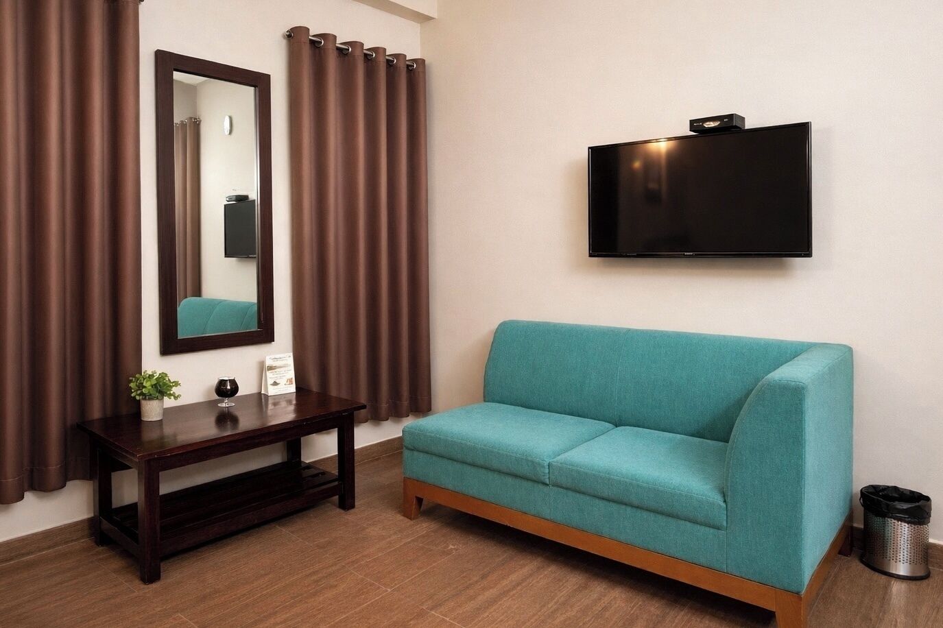 A room with a teal sofa, a wooden console table, a mirror, brown curtains, a TV mounted on the wall, and a trash can.
