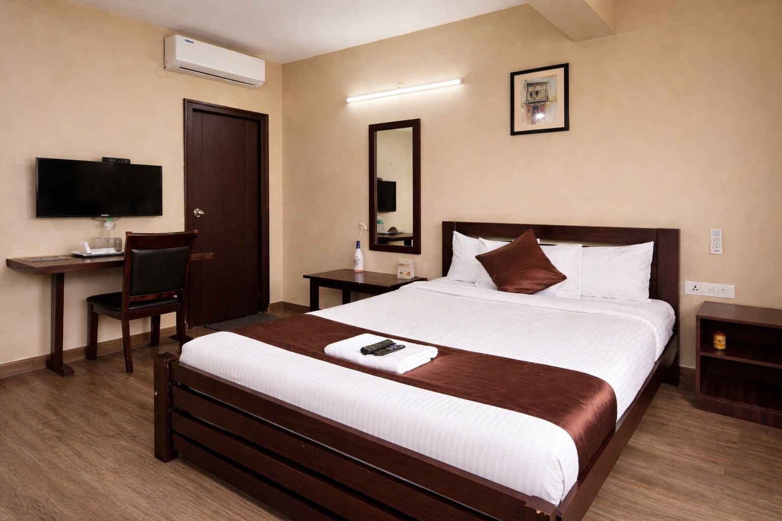 Una habitación de hotel con cama, escritorio, silla, televisor, espejo y mesita de noche.