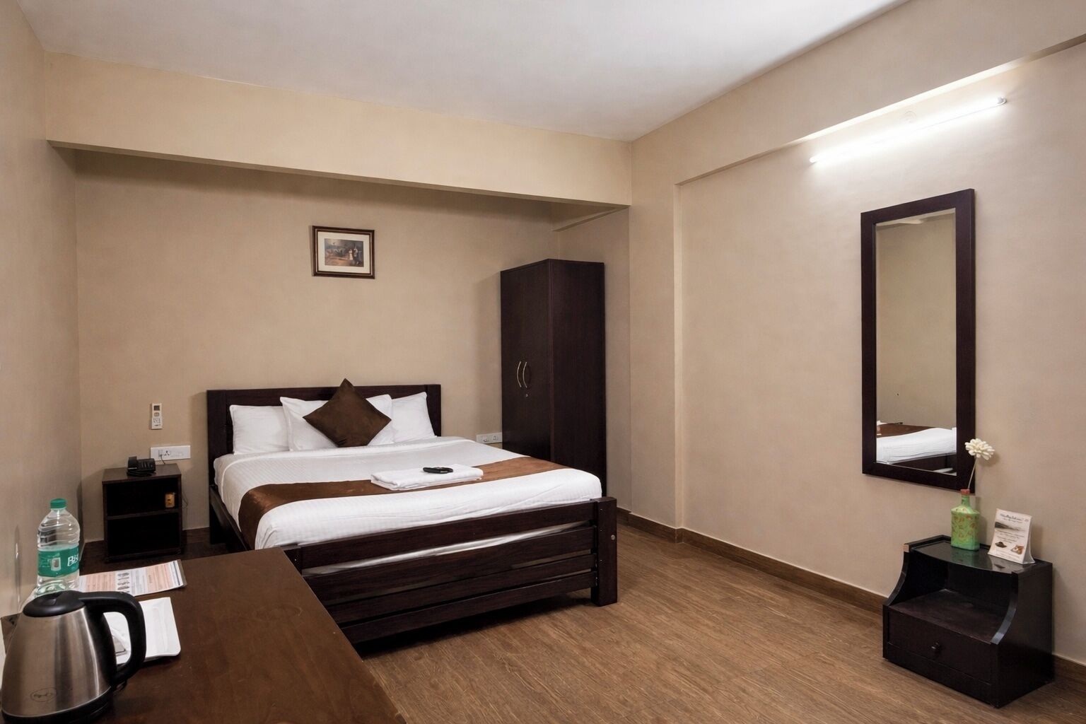 Une chambre d’hôtel comprenant un lit, une table de chevet, un bureau avec une bouilloire, une armoire, un miroir et une petite table avec un vase.