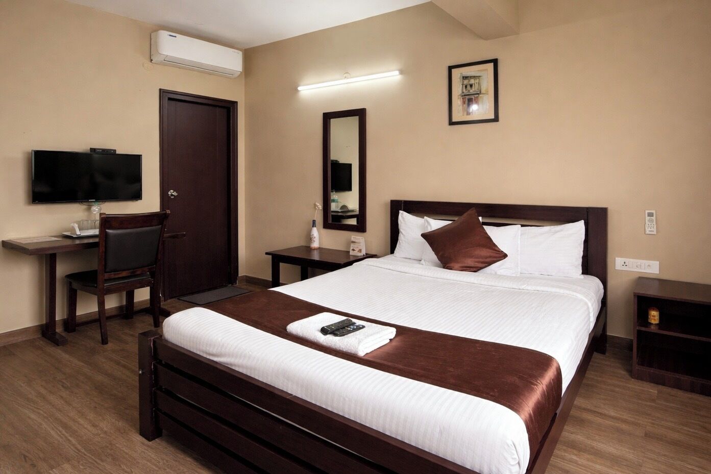 A hotel room with a bed, a desk with a chair, a TV, a bedside table, and a mirror.