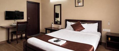 A hotel room with a bed, a desk with a chair, a TV, a bedside table, and a mirror.