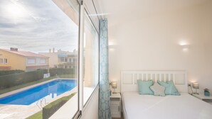 A bedroom with a large window, a bed with pillows, a bedside table with a lamp, and a view of a swimming pool and neighboring buildings.