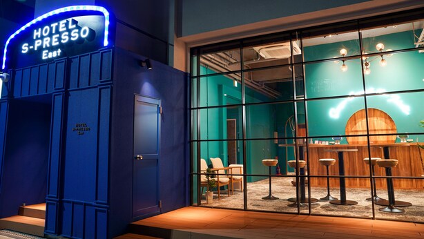 Entrada de hotel com um letreiro de neon, área de recepção com um bar e área de estar com cadeiras e mesas.