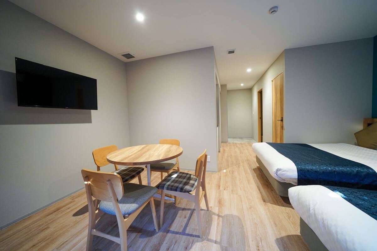 A modern hotel room with a wooden dining table, two chairs, a single bed, and a flat-screen TV mounted on the wall.