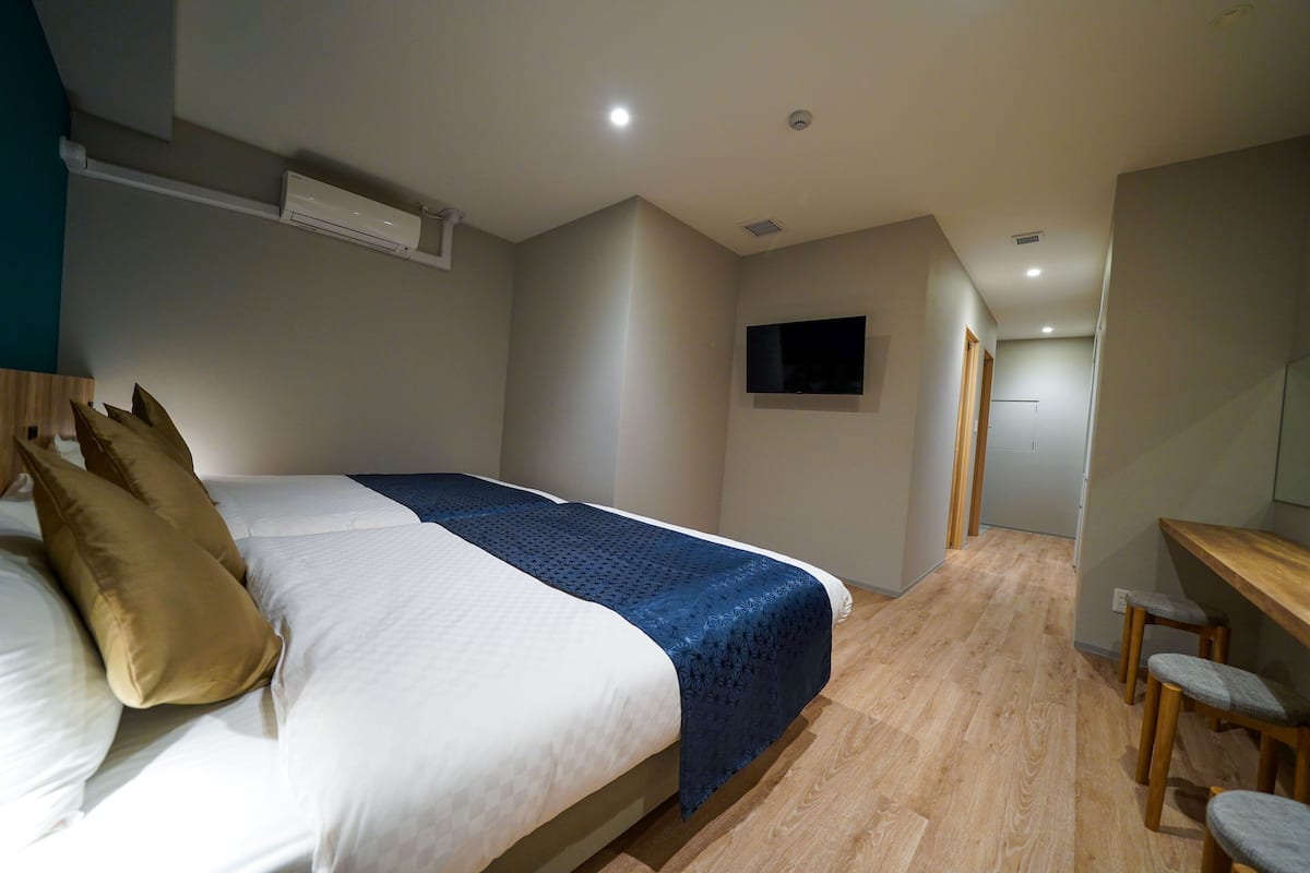 A hotel room with a bed, a TV mounted on the wall, a desk with chairs, and a wooden floor.