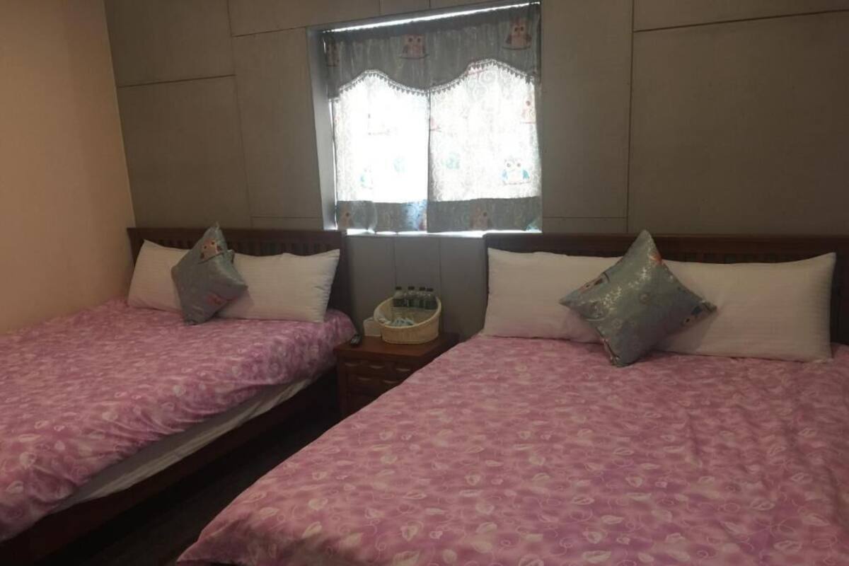 Una habitación con dos camas, cada una con cubrecama y almohadas estampadas con motivos florales rosas. Hay una ventana con cortinas de encaje y una mesita con una cesta entre las camas.
