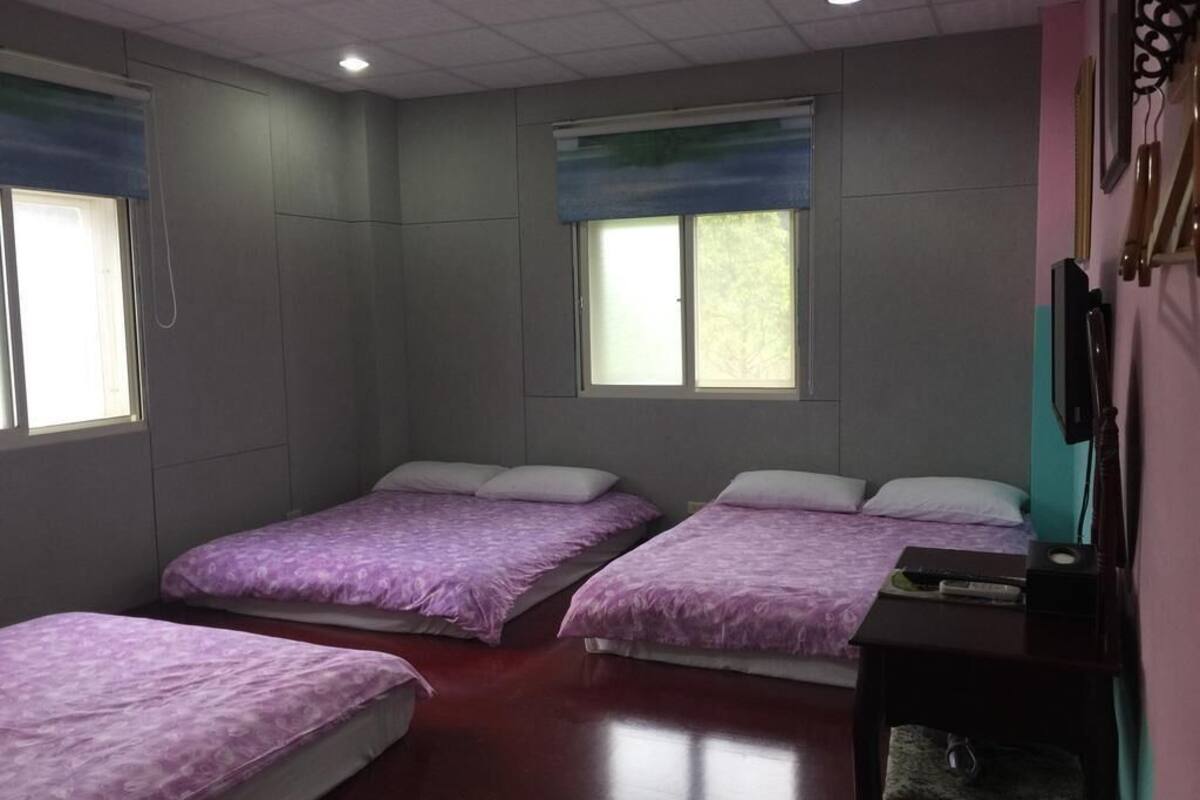 Una habitación con cuatro camas, cada una con cubrecama morado, una mesita con lámpara y una ventana que da a árboles.