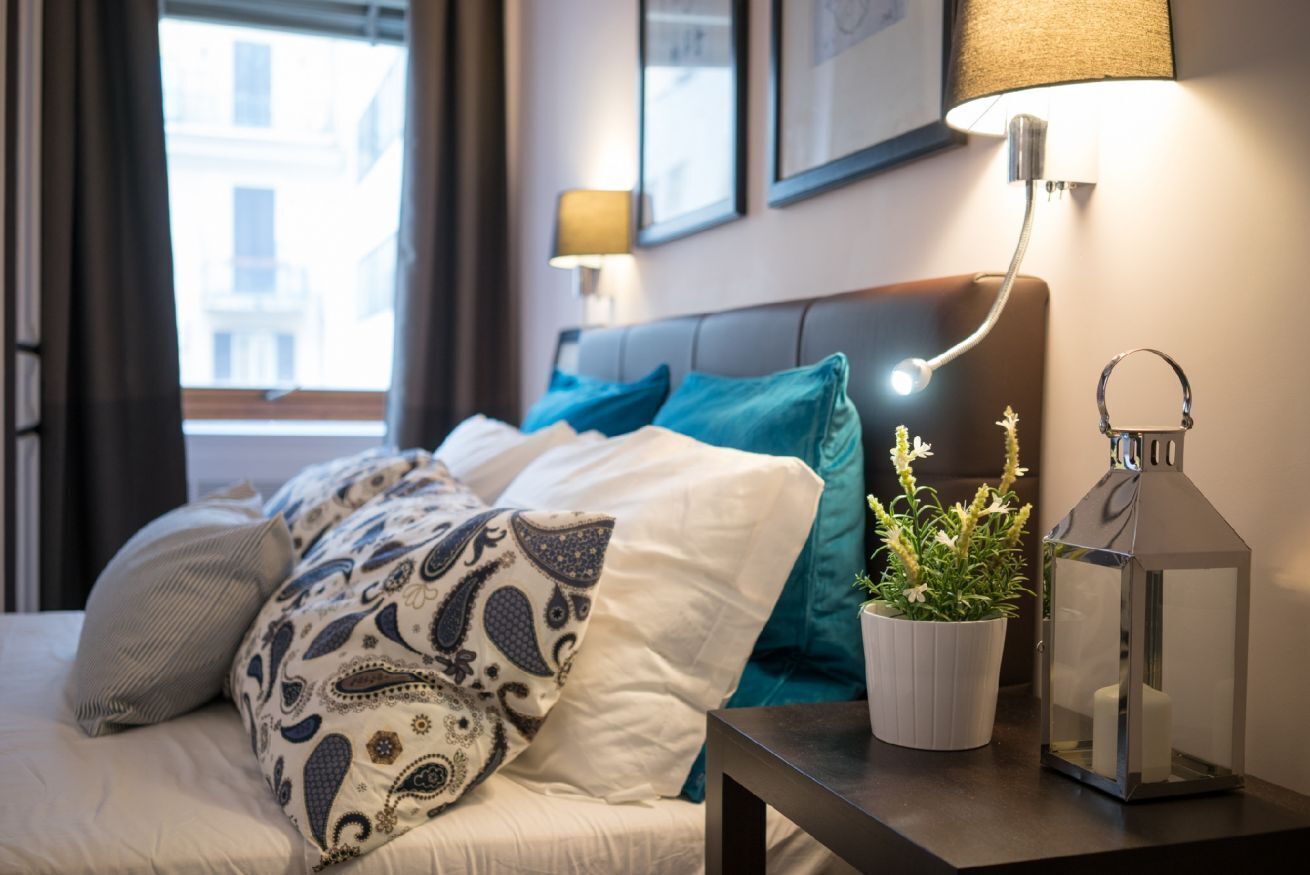 A neatly made bed with patterned pillows, a bedside plant, a lantern, and a wall-mounted lamp.