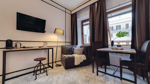 A hotel room with a flat-screen TV, a desk with a lamp, a sofa, a dining table with chairs, and a large window with curtains.