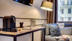 A hotel room with a desk, a coffee maker, a sign indicating WiFi availability, a lamp, a sofa with a patterned pillow, and a television mounted on the wall.