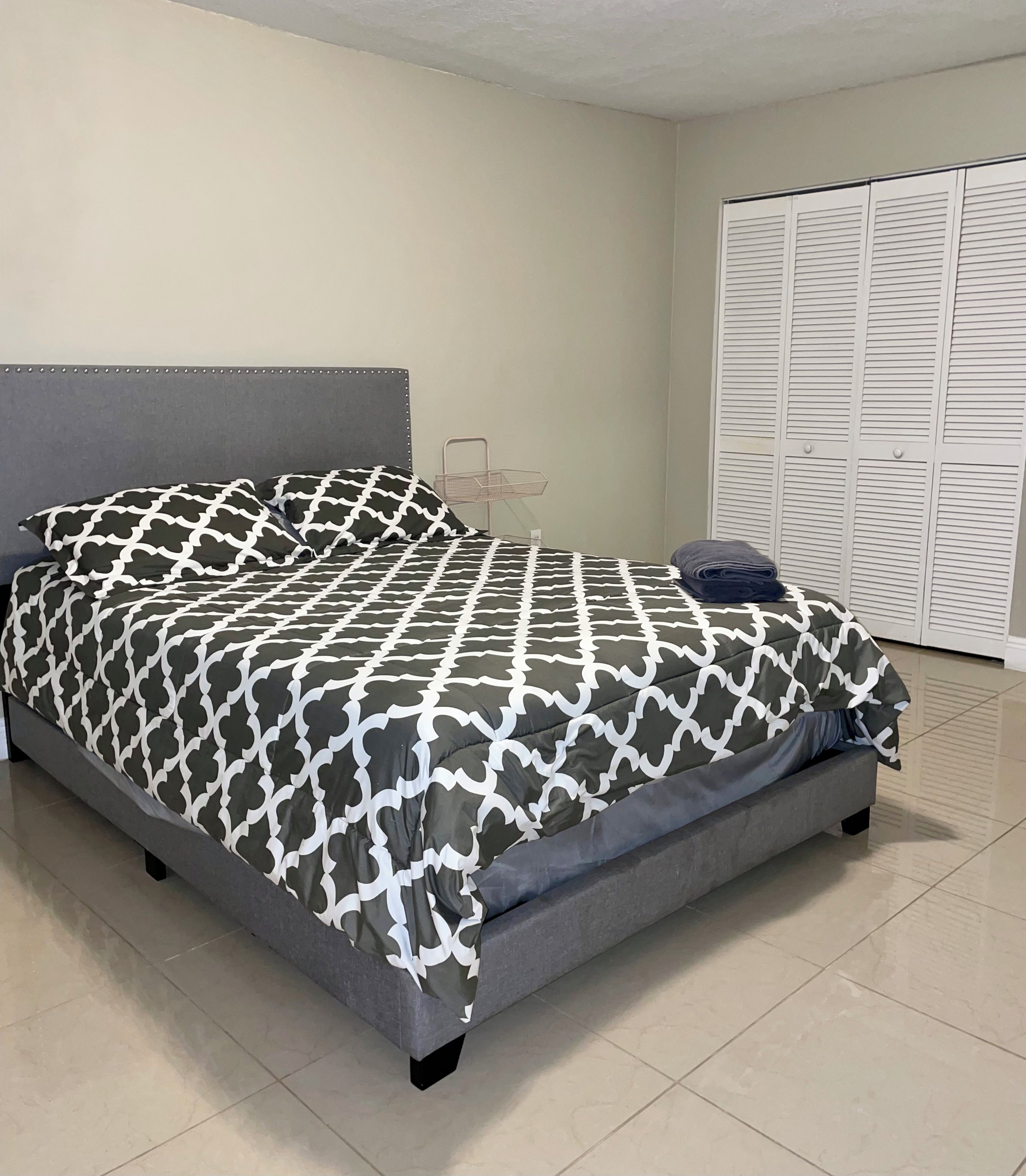 A bed with a patterned bedspread, a grey headboard, and a metal storage unit against the wall.
