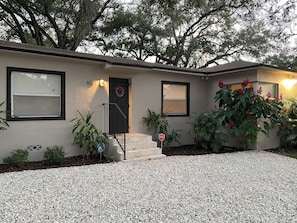 A single-story house with a front door, two windows, a small porch, and a gravel driveway.