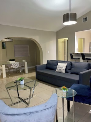 A modern living room with a sofa, a glass coffee table, and a dining area with chairs and a table.